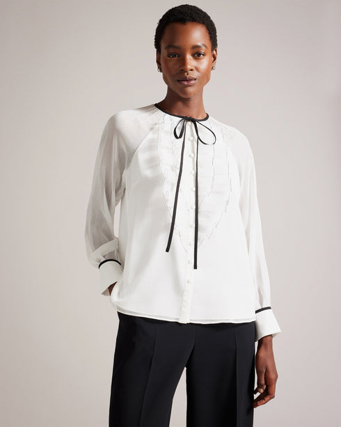 KELEIH-Tops-Channel vintage glamour with a modern twist in the KELEIH blouse. Its delicate organza ruffles and darts exude timeless elegance, while the crew neck keeps it versatile. Whether you're attending a formal event or dressing up for a date night, this blouse will effortlessly elevate your look. Blouse Organza ruffles on front Long sleeves Contrast stitching and neck tie Button fastening Darts on shoulders Crew neck with contrast binding Silvia is 5'11" / 1.8m and wears White in a Ted size 1- Ted Bak